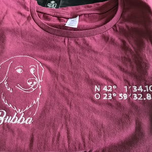 Puede incluir: Camiseta granate con un gr&aacute;fico de perro bordado en blanco y el nombre "Bubba" debajo. Las coordenadas est&aacute;n bordadas en blanco en el lado derecho de la camiseta. La camiseta est&aacute; doblada.
