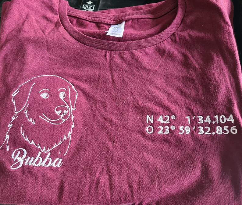 Puede incluir: Camiseta granate con un gr&aacute;fico de perro bordado en blanco y el nombre "Bubba" debajo. Las coordenadas est&aacute;n bordadas en blanco en el lado derecho de la camiseta. La camiseta est&aacute; doblada.