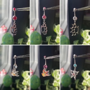 Puede incluir: Colección de piercings para el ombligo con varios diseños. Incluye anillos con un puño americano, un pájaro y una mariposa. Cada anillo tiene una gema en la parte superior e inferior. Los anillos se sostienen con pinzas sobre un fondo de plantas verdes.