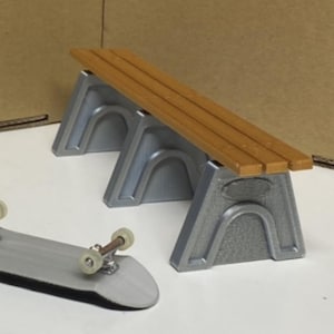 May include: A miniature skateboard and a gray and brown bench set. The skateboard has a gray deck with black grip tape and silver trucks with clear wheels. The bench has a brown top and gray supports.