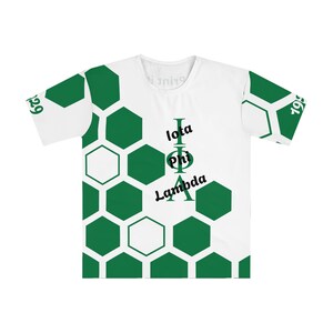 以下が含まれることがあります： 緑色のハニカム模様とギリシャ文字のイオタ、ファイ、ラムダがプリントされた白いTシャツ。袖には「1929」と「1969」の数字がプリントされています。Tシャツはクルーネックです。