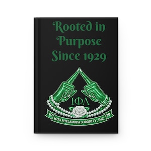 May include: Black journal with the text "Rooted in Purpose Since 1929" in green. The cover features a green emblem with hands forming a triangle, a pearl necklace, and a white rose. The text "IOTA PHI LAMBDA SORORITY, INC." is also present.