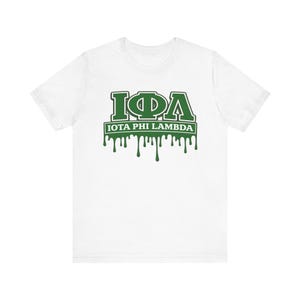 Puede incluir: Camiseta blanca con un gráfico verde. El gráfico presenta las letras griegas Iota, Phi y Lambda, con las palabras "IOTA PHI LAMBDA" debajo. La pintura verde gotea desde la parte inferior del gráfico.