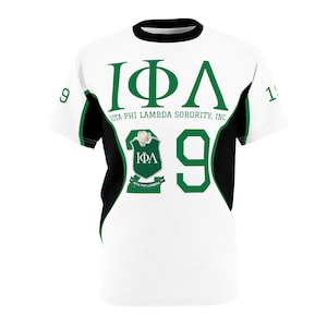 Könnte beinhalten: Weißes T-Shirt mit grünen und schwarzen Akzenten. Das Shirt zeigt die griechischen Buchstaben IΦΛ in Grün, den Text "IOTA PHI LAMBDA SORORITY, INC." und die Zahl "29". Ein Wappen mit einer Blume ist ebenfalls zu sehen.