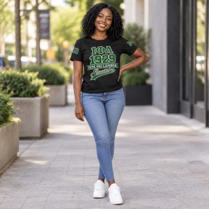 Puede incluir: Una camiseta negra de manga corta con el texto en verde y blanco IOTA PHI LAMBDA 1929 y un logotipo decorativo, combinada con pantalones vaqueros azul claro y zapatillas blancas. Las mangas de la camiseta también muestran el número 1929 en verde.