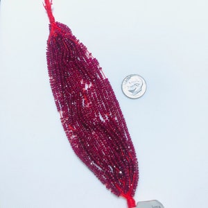 May include: A strand of small, faceted, deep red ruby beads strung on red thread. A US dime coin is placed next to the beads for scale. A white paper tag at the top displays the text "Gw 280.75", "Nw 279.75", and "L-37". A tag at the bottom reads "Ruby".