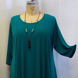 May include: Teal blue, asymmetrical, long-sleeved tunic top with a round neckline. The top is worn on a mannequin and features a brown wooden necklace with turquoise beads.