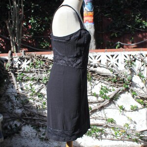 May include: A black dress with spaghetti straps and a fitted bodice. The dress features lace detailing around the bust, waist, and hem. The dress is displayed on a mannequin in front of a white wall with vines.