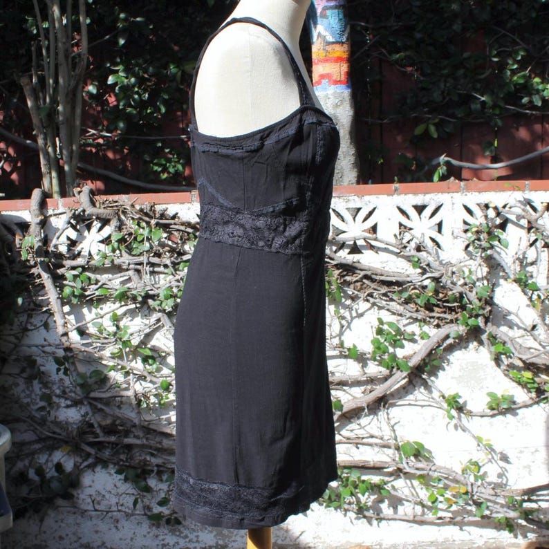 May include: A black dress with spaghetti straps and a fitted bodice. The dress features lace detailing around the bust, waist, and hem. The dress is displayed on a mannequin in front of a white wall with vines.