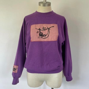 May include: Purple long-sleeved sweatshirt with a red and white gingham patch featuring a black and white illustration of a bird on a branch. The sweatshirt also has a smaller gingham patch on the left sleeve.