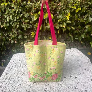 May include: A green quilted tote bag with pink floral print and pink handles. The bag is sitting on a white crocheted surface.