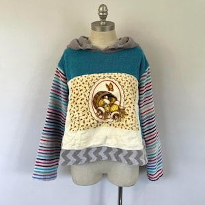 May include: A teal blue, white, and gray patchwork hoodie with a striped sleeve. The front of the hoodie features a white patch with a floral pattern and a picture of a basket of fruit with a butterfly.