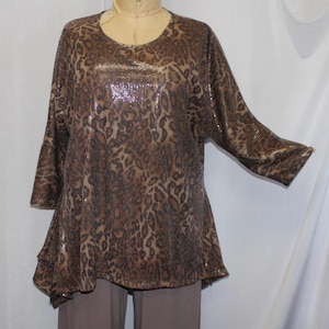 Puede incluir: Una blusa de lentejuelas marrón y dorada con estampado de leopardo. La blusa tiene un escote redondo y mangas tres cuartos. La blusa es asimétrica y cae en punta en la parte delantera.