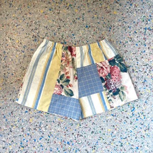 May include: A patchwork skirt featuring various floral and striped patterns in shades of blue, yellow, and white. The skirt has an elastic waistband and a mix of fabrics, creating a unique, handcrafted look. The skirt is a mini-skirt.