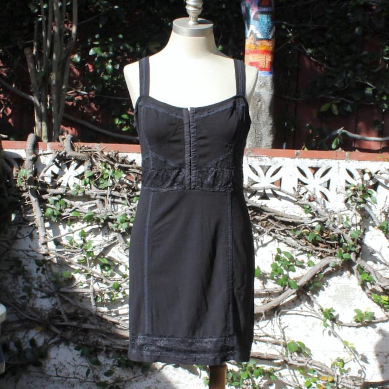 May include: A black dress with spaghetti straps and a fitted bodice. The dress features lace detailing across the chest and hemline, with vertical seams adding structure. The dress is displayed on a mannequin.
