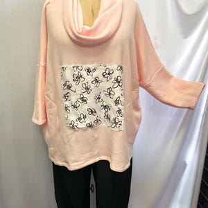 Może przedstawiać: Różowa bluza z kapturem i dekoltem typu cowl neck z białą kieszenią z czarnym nadrukiem kwiatowym. Bluza jest połączona z czarnymi spodniami.