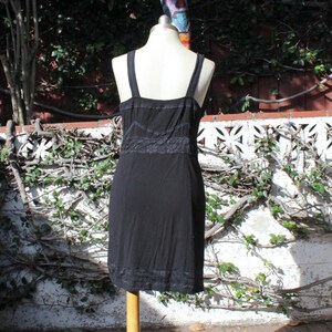 May include: A black dress with thin straps and a fitted bodice. The dress features a lace detail at the waist and hem. The dress is displayed on a mannequin in an outdoor setting with a white wall and greenery in the background.
