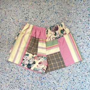 May include: Patchwork shorts featuring a mix of floral, plaid, and striped patterns in shades of yellow, pink, and brown. The shorts have an elastic waistband and a unique, handcrafted appearance.