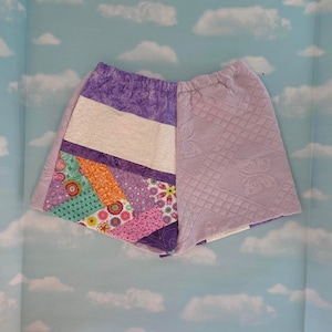 May include: Purple and white patchwork shorts with a quilted design. The shorts are made of different fabrics and have a variety of floral patterns.