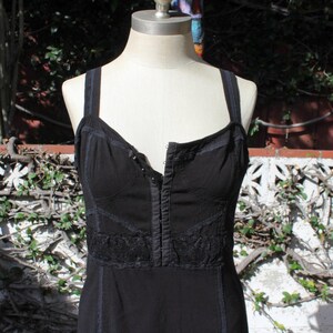 May include: Black dress with a fitted bodice and spaghetti straps. The dress features a lace detail across the midsection and a button-up front. The dress is displayed on a mannequin against a backdrop of greenery and a white wall.