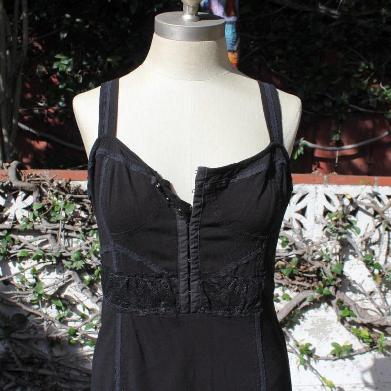 May include: Black dress with a fitted bodice and spaghetti straps. The dress features a lace detail across the midsection and a button-up front. The dress is displayed on a mannequin against a backdrop of greenery and a white wall.