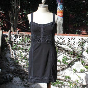 May include: Black dress with spaghetti straps, displayed on a mannequin. The dress features a fitted bodice with decorative stitching and lace detailing. The skirt is a simple A-line shape, and the hemline is trimmed with lace. The dress is made of a dark fabric.