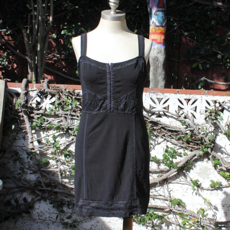 May include: Black dress with spaghetti straps, displayed on a mannequin. The dress features a fitted bodice with decorative stitching and lace detailing. The skirt is a simple A-line shape, and the hemline is trimmed with lace. The dress is made of a dark fabric.