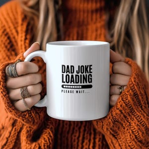 May include: A white ceramic mug featuring the black printed text DAD JOKE LOADING above a horizontal progress bar graphic and the text PLEASE WAIT. The mug has a smooth cylindrical body and a curved handle on the left side for easy gripping.