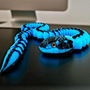 May include: A blue and black toy snake is coiled on a dark surface. The snake has a segmented body with black stripes and a detailed head with orange eyes. The toy is a vibrant, eye-catching design.