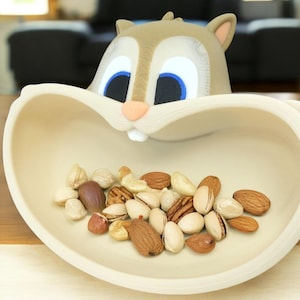 May include: A whimsical, off-white squirrel-shaped bowl filled with a variety of nuts. The squirrel has tan fur, blue eyes, and a pink nose. The bowl is on a wooden surface and is filled with almonds, pecans, and other nuts.