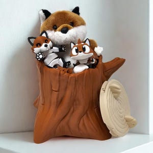 May include: A brown tree stump with a brown, white, and black plush fox and two smaller brown, white, and black fox figurines inside. The stump has a light brown, circular lid.