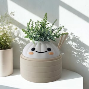 May include: A whimsical, beige and white planter shaped like a dumpling, with a smiling face and small green plant inside. The planter has a handle and is sitting on a white surface.