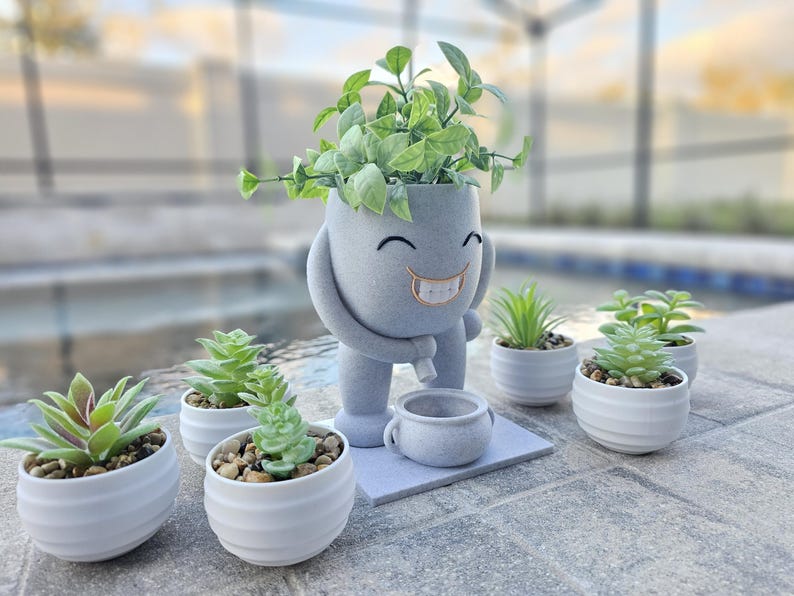 A quirky plant pot with a smiling face and a playful pose is placed outdoors by a pool, surrounded by small white pots with vibrant green succulents. The planter features a modern, speckled gray finish and a built-in saucer for drainage. The bright greenery contrasts with the neutral tones, making it a perfect decorative piece for patios, balconies, or indoor spaces. The relaxed setting highlights the pot’s fun design, adding charm to any plant collection.