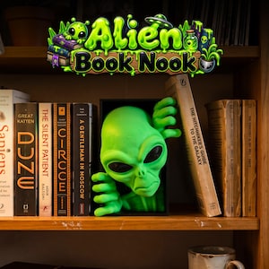 May include: A bright green alien figure emerging from a black rectangular book nook insert, positioned between several books on a wooden shelf. The alien has large black eyes and hands reaching out. The text Alien Book Nook is displayed above.