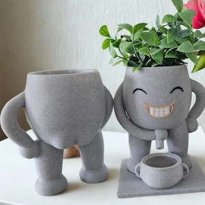 May include: Two gray, anthropomorphic planters with arms and legs. One planter has a smiling face with a gold-colored mouth and holds a small cup. The other planter is plain. Green plants are in the smiling planter.