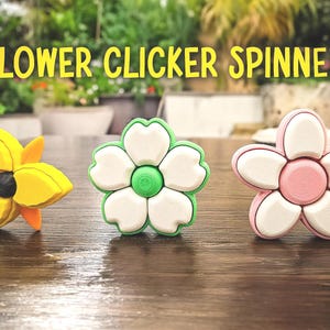 May include: Three flower-shaped fidget spinners in yellow, white and green, and pink and white. The text "FLOWER CLICKER SPINNER" is in yellow above the spinners. The spinners are on a brown wooden surface.