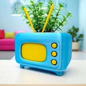 May include: A blue and yellow planter shaped like a retro television set. The planter has a yellow screen and three yellow buttons. Two yellow antennas extend from the top, and the planter is filled with green plants.