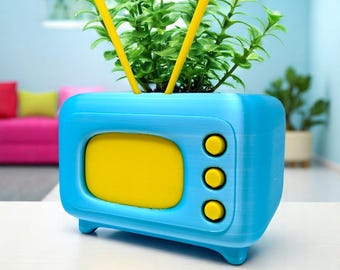 May include: A blue and yellow planter shaped like a retro television set. The planter has a yellow screen and three yellow buttons. Two yellow antennas extend from the top, and the planter is filled with green plants.
