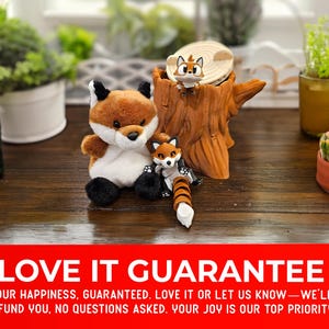 A charming setup featuring a plush fox sitting beside a tree stump container with two articulated fox figurines playfully posed around it. The cozy scene is decorated with potted plants in the background on a wooden surface. A bold red banner along the bottom highlights a "Love It Guarantee" message, promising customer satisfaction or a full refund.