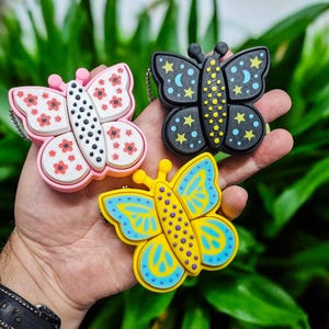 May include: Three butterfly-shaped keychains in pink, black, and yellow. The pink butterfly has red flower accents, the black one has moon and star designs, and the yellow one has blue details. Each keychain has a silver chain.