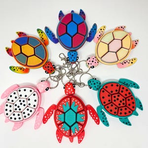 May include: A collection of colorful turtle keychains. Each keychain features a turtle design with a unique color scheme, including blue, red, pink, yellow, and green. The turtles have black dot patterns and are attached to silver keyrings.