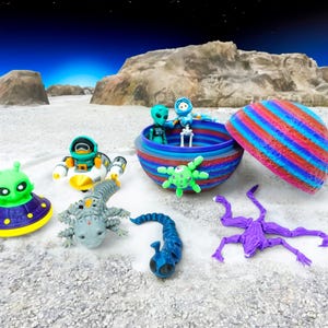 May include: A collection of colorful alien figurines and a striped container. The figurines include a green alien in a UFO, a robot, a gray lizard-like creature, a blue alien, a green star-shaped alien, and a purple creature.