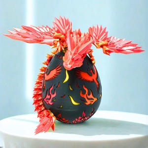 May include: A black, egg-shaped decorative object with red and yellow flame cutouts and phoenix designs. The top features a red, origami-style dragon with outstretched wings. The object is on a white surface against a light blue background.