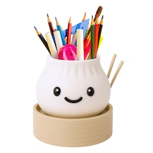 May include: A white, dumpling-shaped desk organizer with a smiling face, holding pencils, pens, and art supplies. The organizer sits on a beige, circular base and includes two wooden chopsticks. A fun and functional desk accessory.