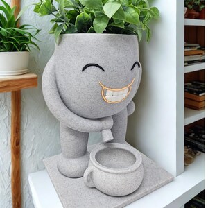 Puede incluir: Una maceta gris sonriente con un dise&ntilde;o caprichoso, que sostiene una taza peque&ntilde;a. La maceta tiene una cara con detalles bordados y est&aacute; llena de plantas verdes y frondosas. La maceta est&aacute; sobre una base cuadrada.