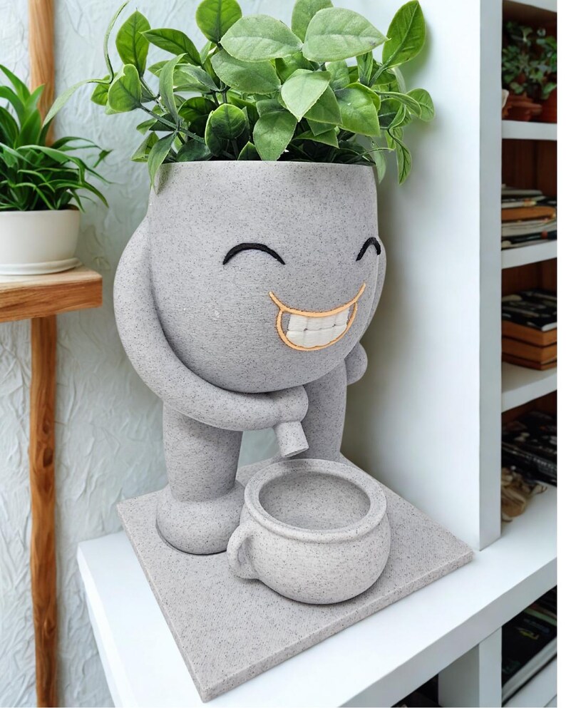 Puede incluir: Una maceta gris sonriente con un dise&ntilde;o caprichoso, que sostiene una taza peque&ntilde;a. La maceta tiene una cara con detalles bordados y est&aacute; llena de plantas verdes y frondosas. La maceta est&aacute; sobre una base cuadrada.