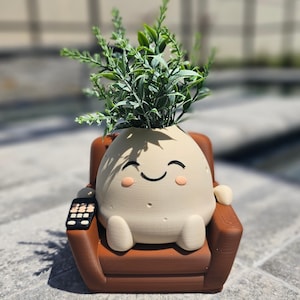 May include: A beige ceramic planter shaped like a smiling potato sitting in a brown armchair with a remote control. The planter has a green leafy plant growing out of the top.