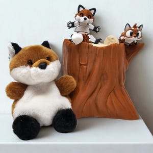May include: A brown plush fox toy sitting next to a brown tree stump. Two smaller fox figurines are perched on top of the stump.