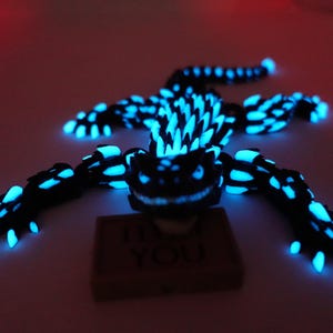 May include: A black and blue glowing lizard figurine with a detailed, textured design. The lizard is lying on a surface, with the text "I <3 YOU" on a small block beneath it. The figurine has glowing blue accents.