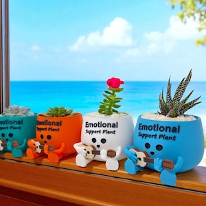 May include: Four colorful ceramic planters with the text "Emotional Support Plant" are displayed on a wooden windowsill. Each planter holds a different succulent or flowering plant. Each planter has a small figure playing a guitar.